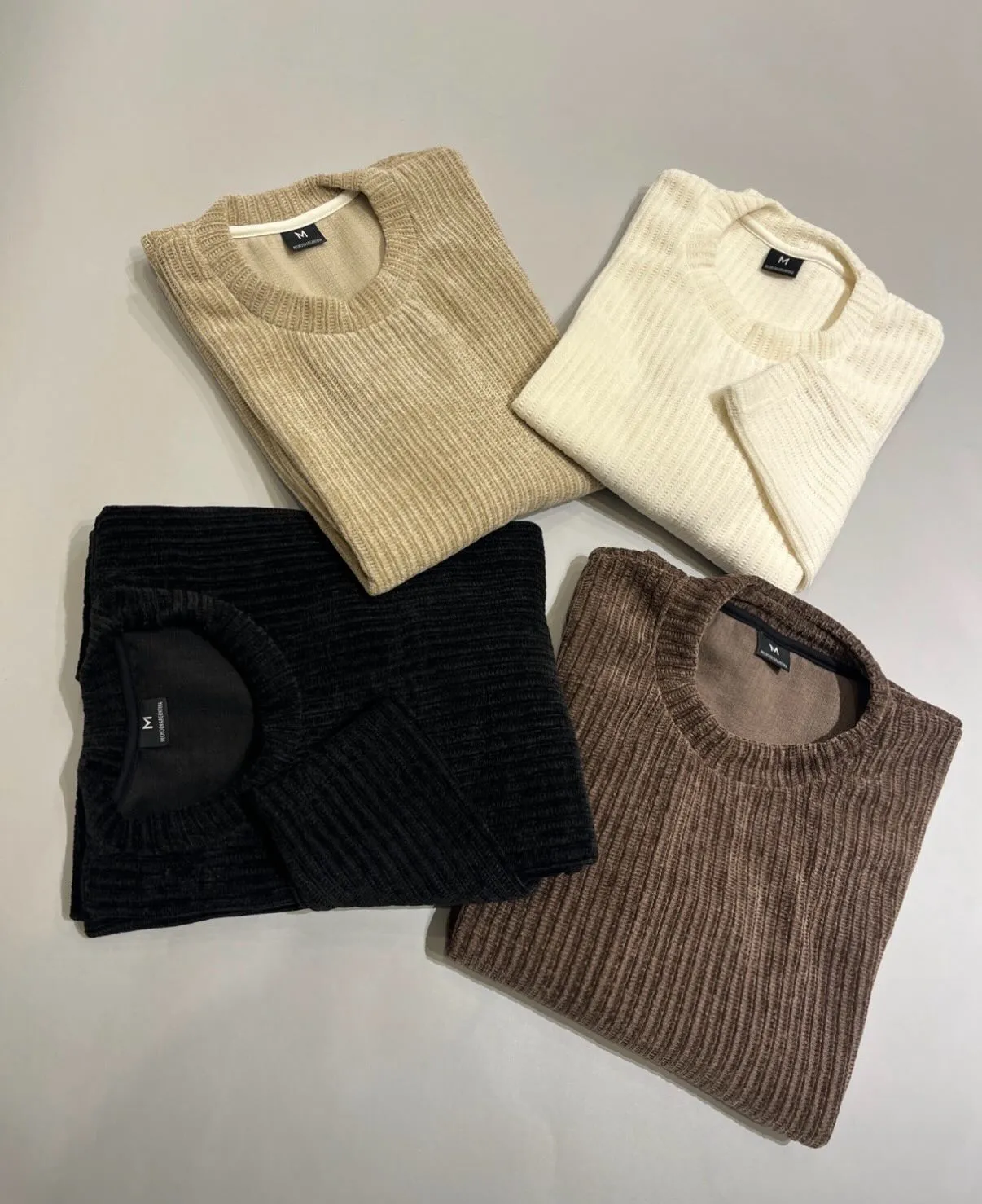 Cuatro sweaters de punto plegados en beige, crudo, negro y marrón sobre fondo blanco